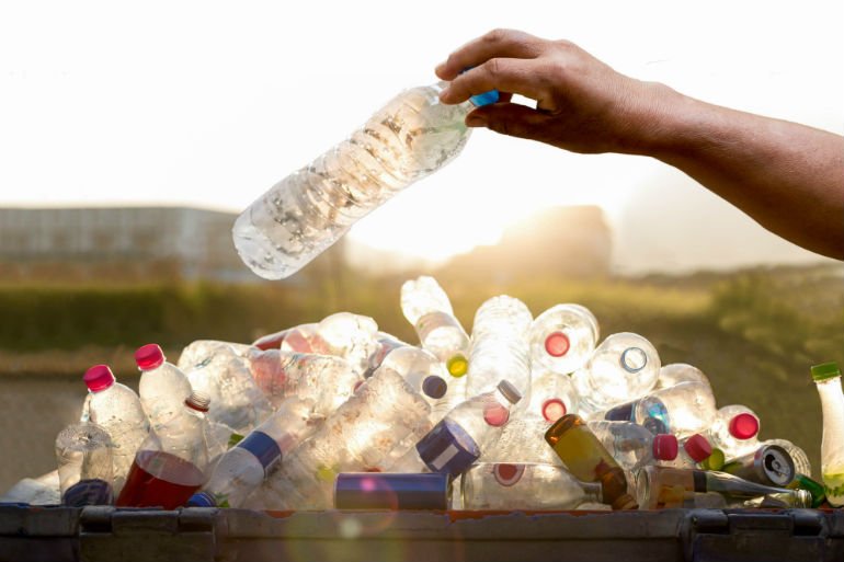 En hand släpper en plastflaska över en hög med återvunna plastflaskor i solljus.