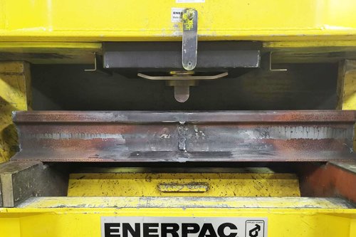 Gul hydraulisk press som böjer en räl i stål, så kallad böjprovning av räl som en metod inom förstörande provning, för att verifiera att rälen uppfyller ställda hållfasthetskrav.