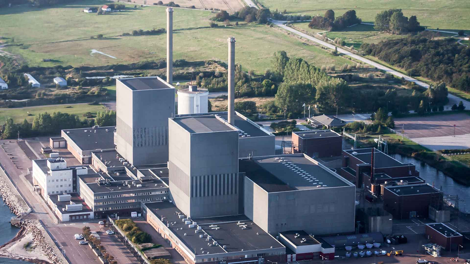 Flygvy över ett stort kärnkraftverk med flera höga skorstenar och industriella byggnader vid kusten, omgiven av grönska och vägar.