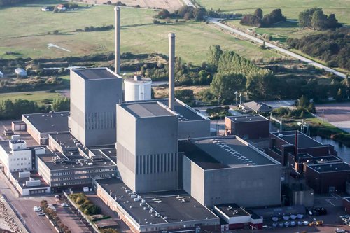 Flygvy över ett stort kärnkraftverk med flera höga skorstenar och industriella byggnader vid kusten, omgiven av grönska och vägar.