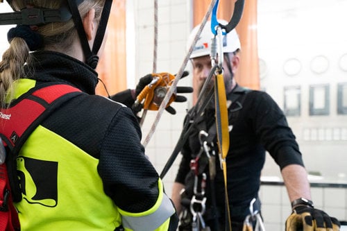 Två personer i skyddsutrustning tränar på arbetssäkerhet med rep och selar inomhus.