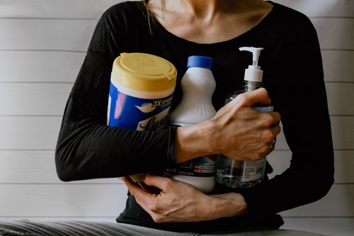 Person holding cleaning supplies including disinfectant wipes, bleach bottle, and hand sanitizer against a white paneled wall background