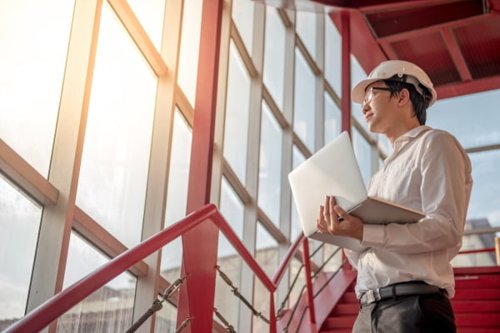 Kiwa engineer wearing a hard hat holding a laptop, standing on red stairs with sunlight streaming through large windows