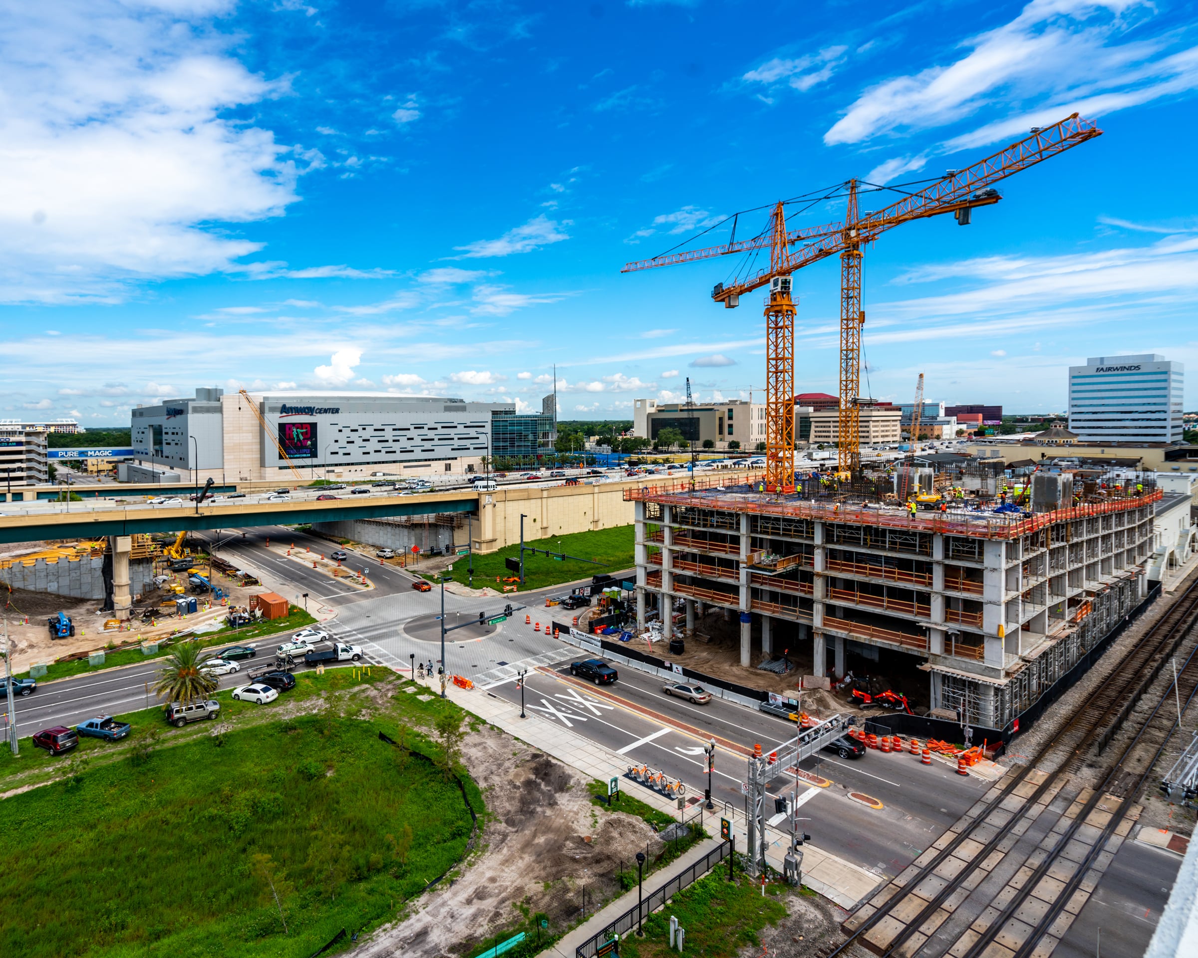 Urban construction site with two tower cranes, a partially built high-rise, and surrounding city roads near a large arena under a blue sky.